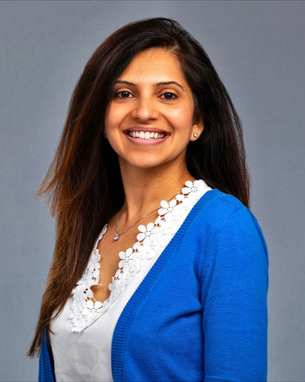 A portrait of Kruti Shah smiling. A woman with brown hair and highlights a light brown complexion wearing a white shirt and blue cardigan.