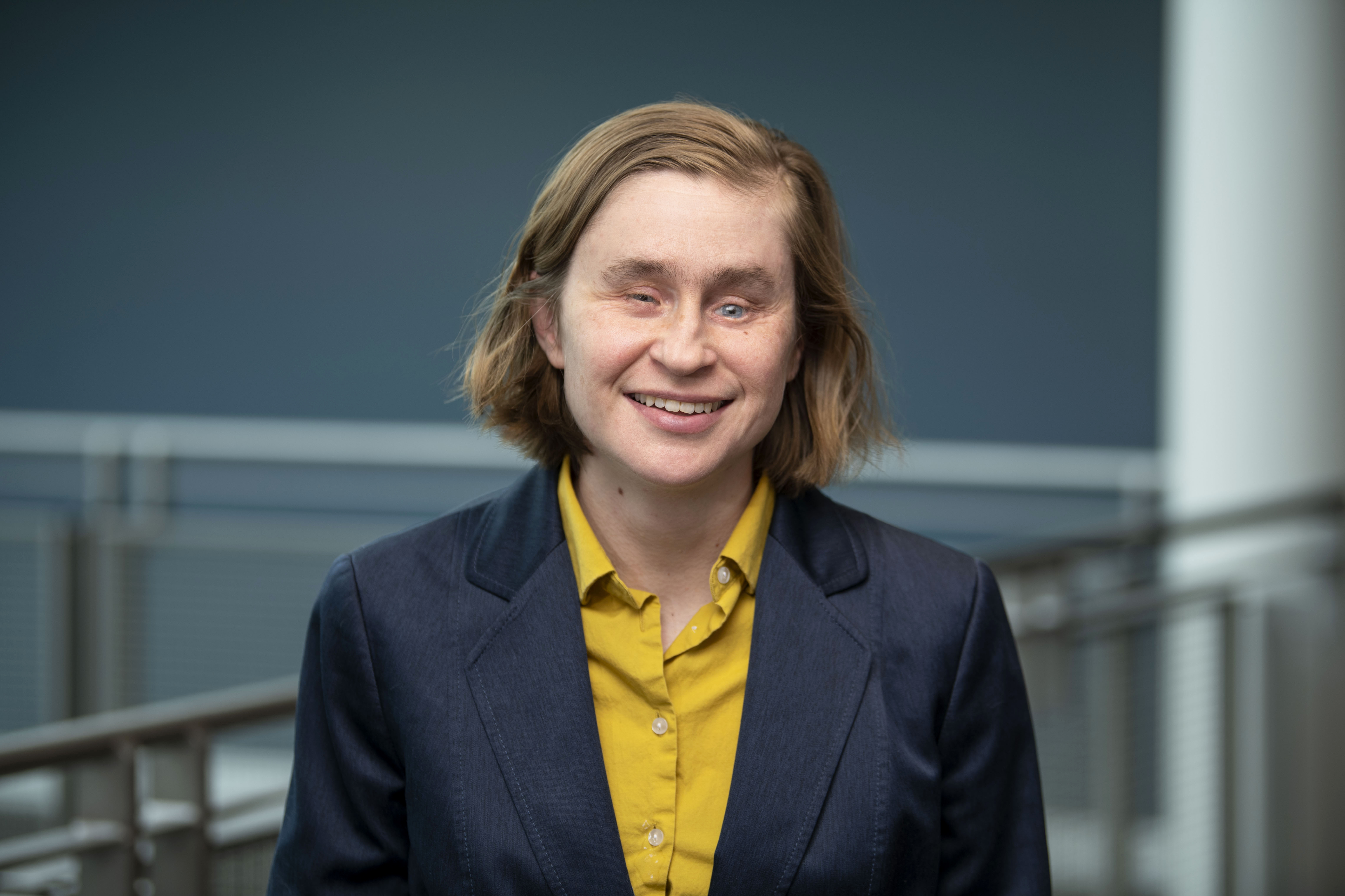 A woman with light brown hair and light to pale skin smiles at the camera, wearing a black blazer and orange button down shirt