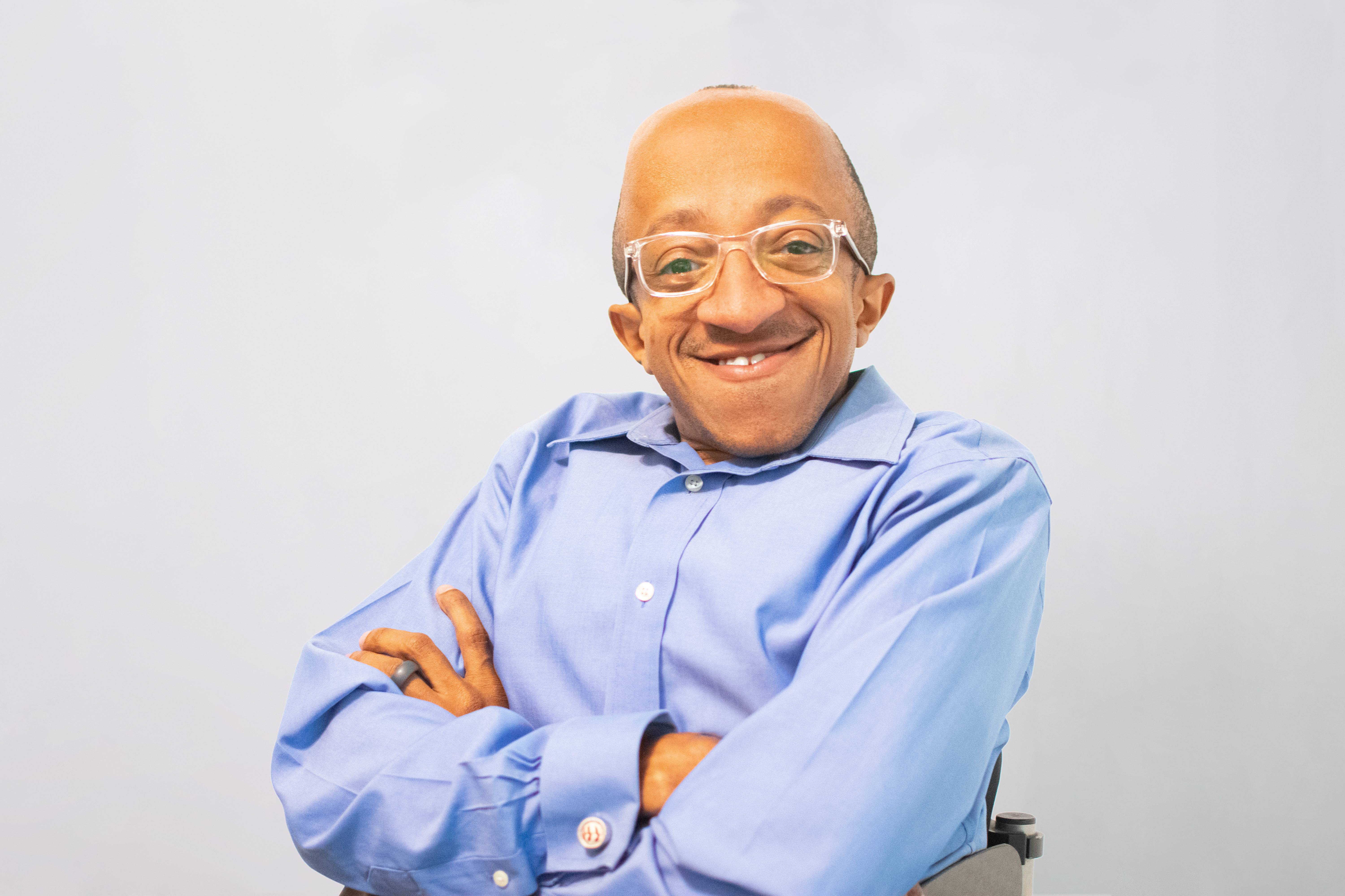 A man with light brown skin wearing a bright blue button-downs shirt and silver framed glasses is smiling at the camera with his arms folded across his chest.