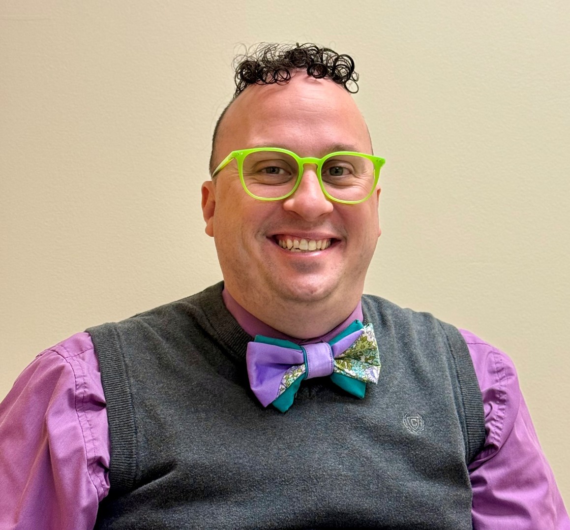 A man with a light to pale skin tone wearing light gray famed glasses, a lavender button-down shirt with a dark gray sweater and a blue and purple bowtie. He is smiling at the camera and has short dark curly hair at the top of his head.