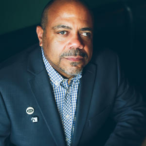 A Black man with medium to to dark skin tone wearing a navy-blue blazer and checkered blue shirt. On the lapel of the blazer are two pins. 