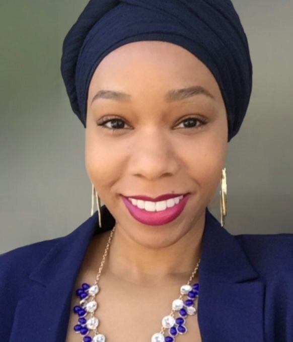 Tayler is smiling in front of a gray background. She is an African American woman wearing a navy headscarf, gold dangle earrings, a blue and silver necklace and a navy blazer top.