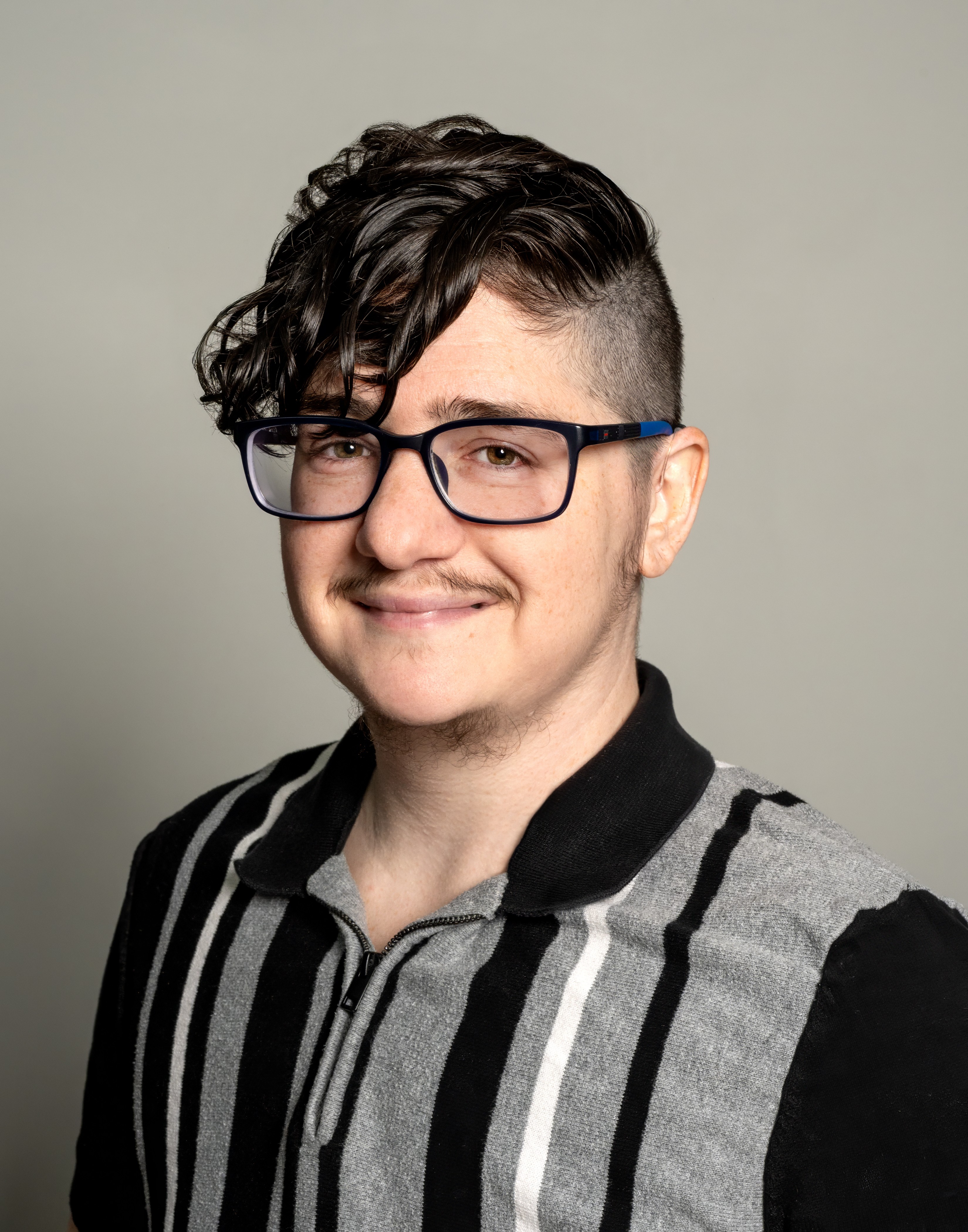 Asher is a white person with a light to pale skin tone wearing a long sleeved, black and vertical gray striped shirt. Asher is wearing black framed glasses and has black wavy hair with shaved sides. 