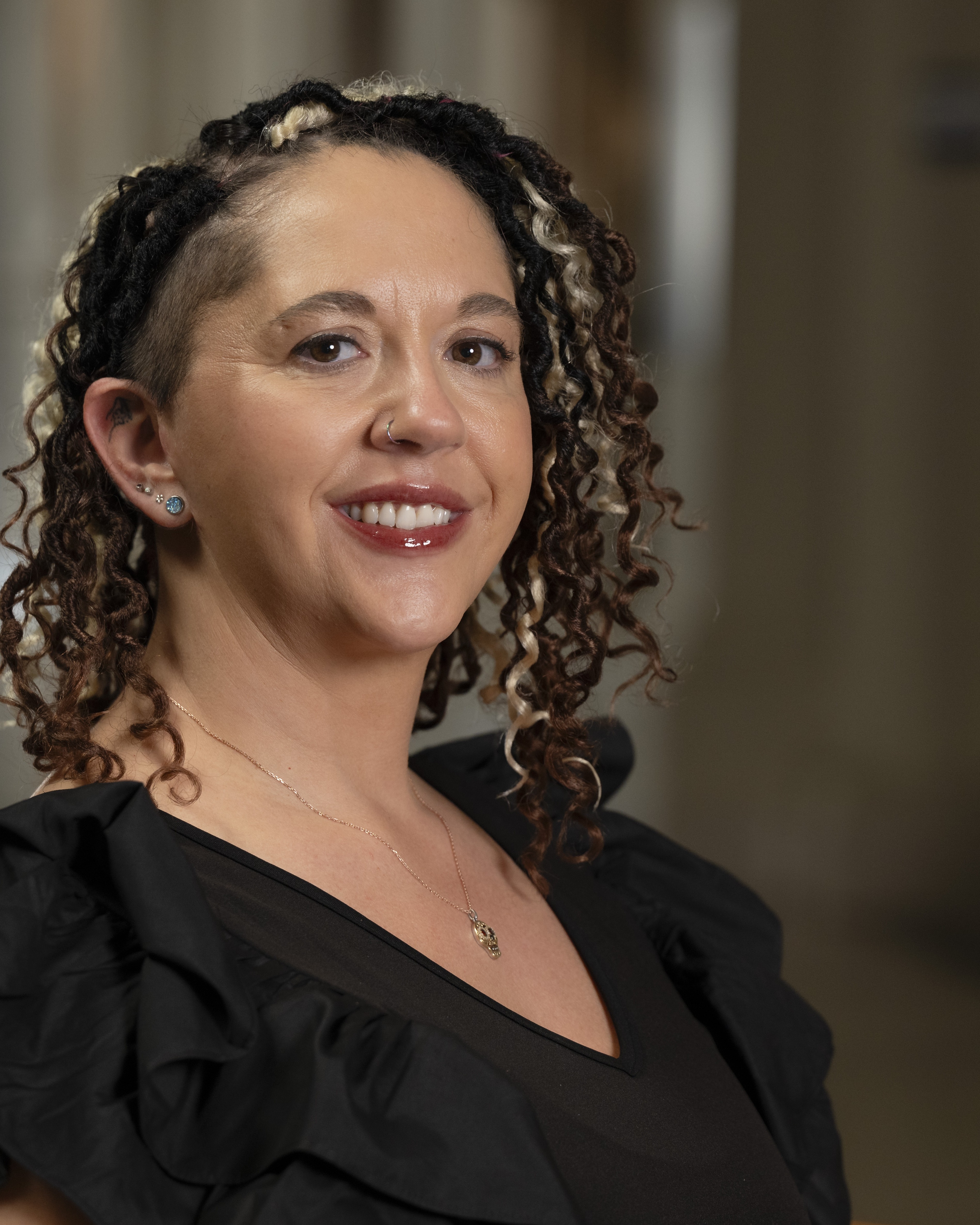 Angelica has light to medium skin tone and chin-length curly brown hair with blonde highlights. Angelica is smiling and is wearing a v-neck back top with ruffles.