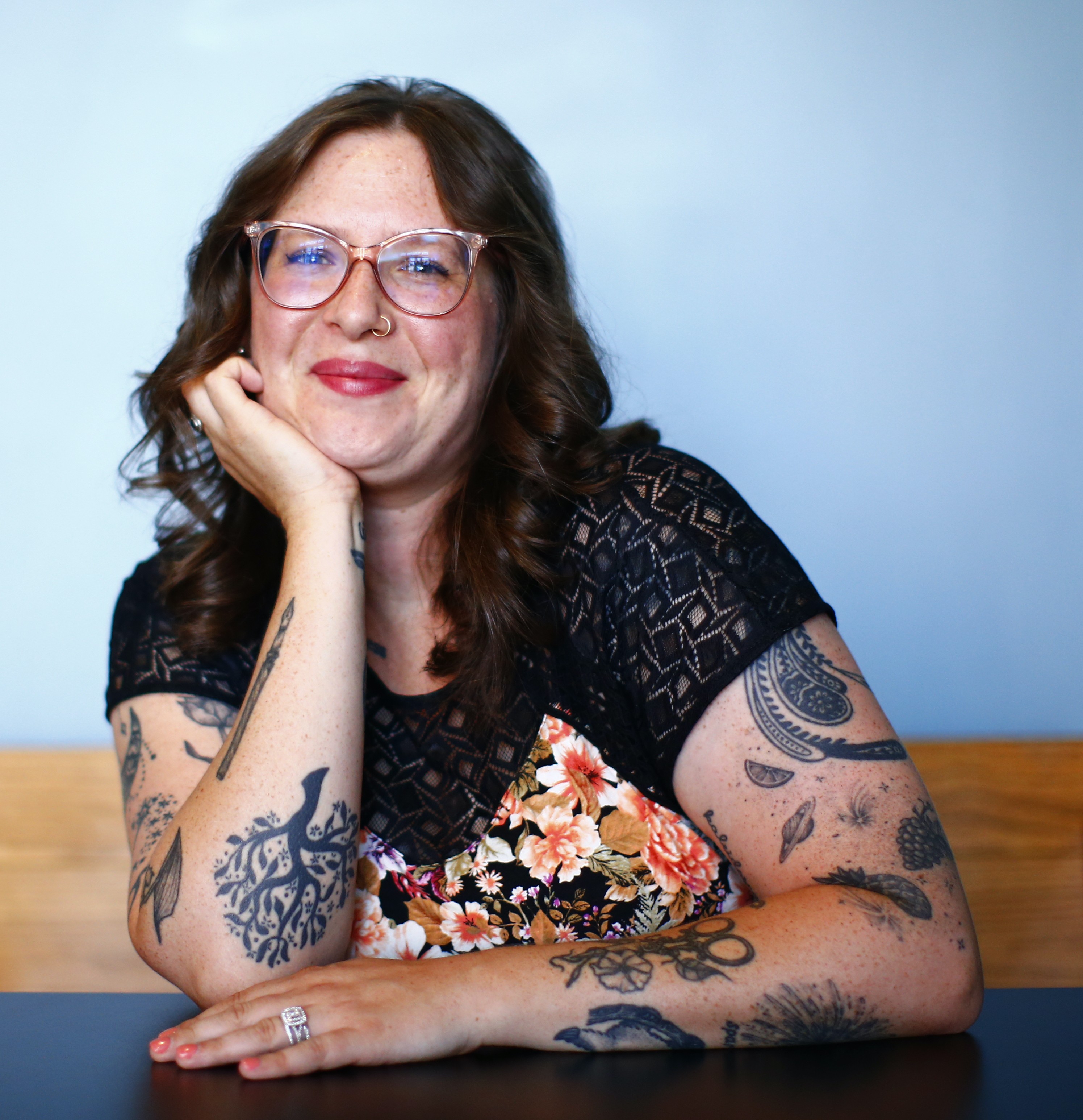 Rachel is woman with light to pale skin, chest-length wavy brown hair and has tattoos on her arms. She is wearing a black and floral top and clear frame glasses.