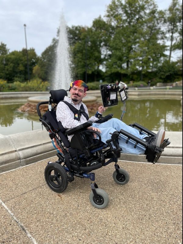 Mark is a man with light to pale skin and a power chair user. He is smiling brightly with his chair cocked upwards, so his legs are at an angle. he is wearing a white shirt and light blue pants.