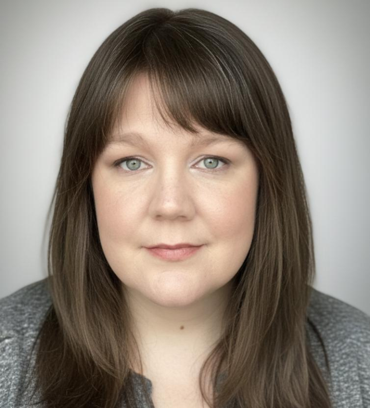 Jenn Jones is smiling against a neutral background. She is a white woman with straight, brown hair cut in bangs. She wears a gray jacket and a blue blouse. 