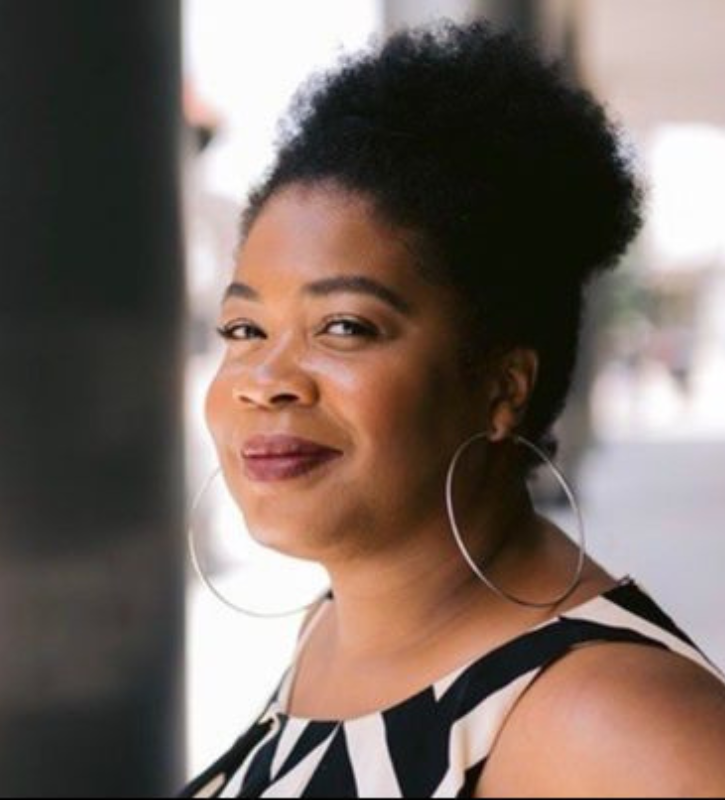 A white woman with curly, blond hair, a pink blazer, and black pants. She is using a manual wheelchair.