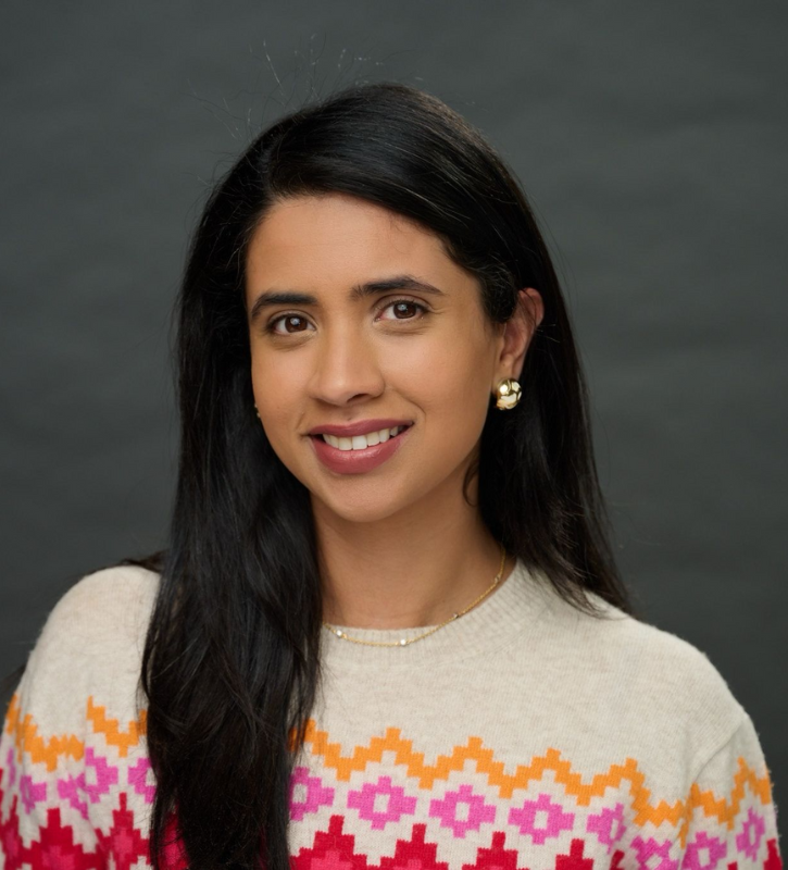 A Brown woman with long, dark hair wearing chunky gold earrings and a red, yellow, cream, and pink top.