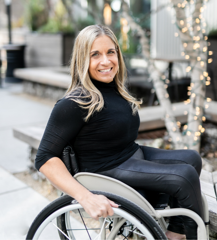 A white woman with straight blond hair and a long-sleeved black shirt. She is using a manual wheelchair.