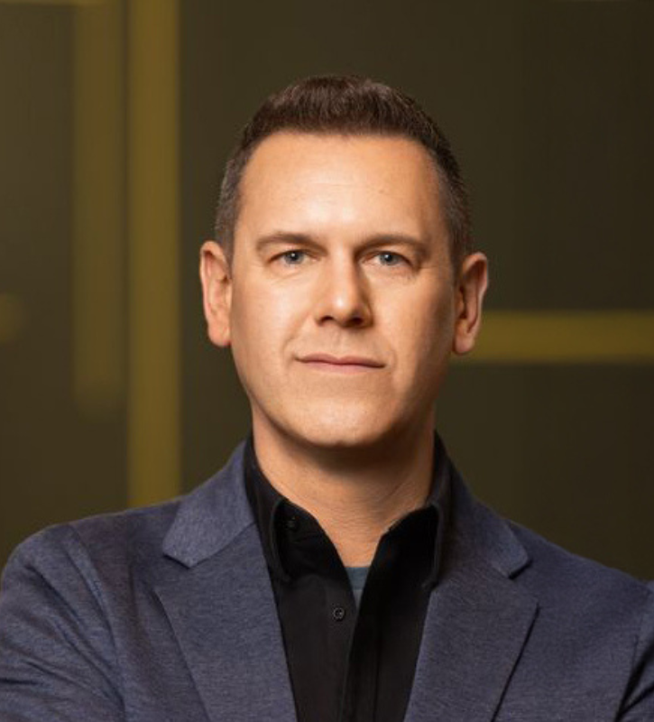 A photo of a White man with short brown hair, wearing a blazer on top of a black sweater.