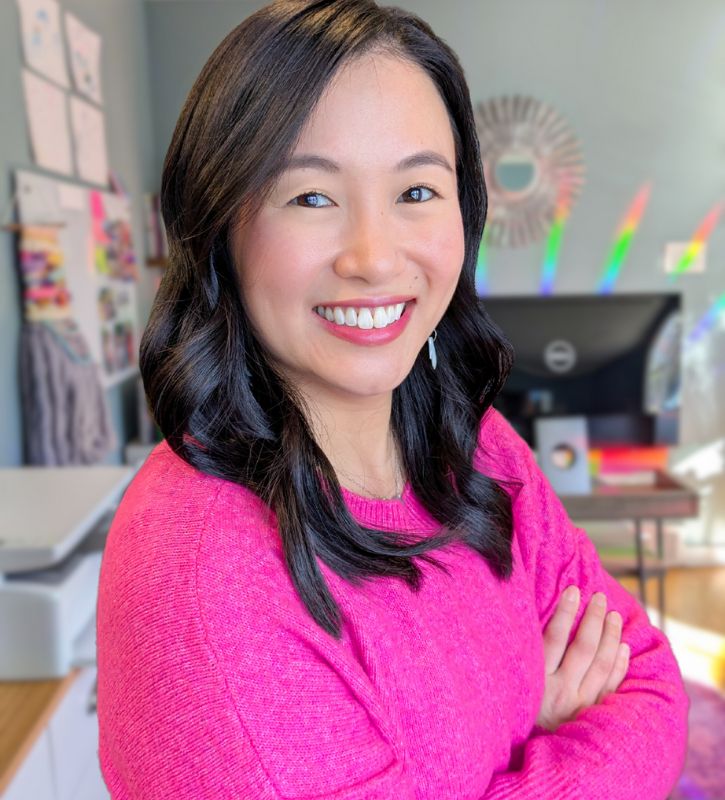 Photo of an Asian woman with long black hair smiling with her arms folded across her chest, wearing a hot pink sweater