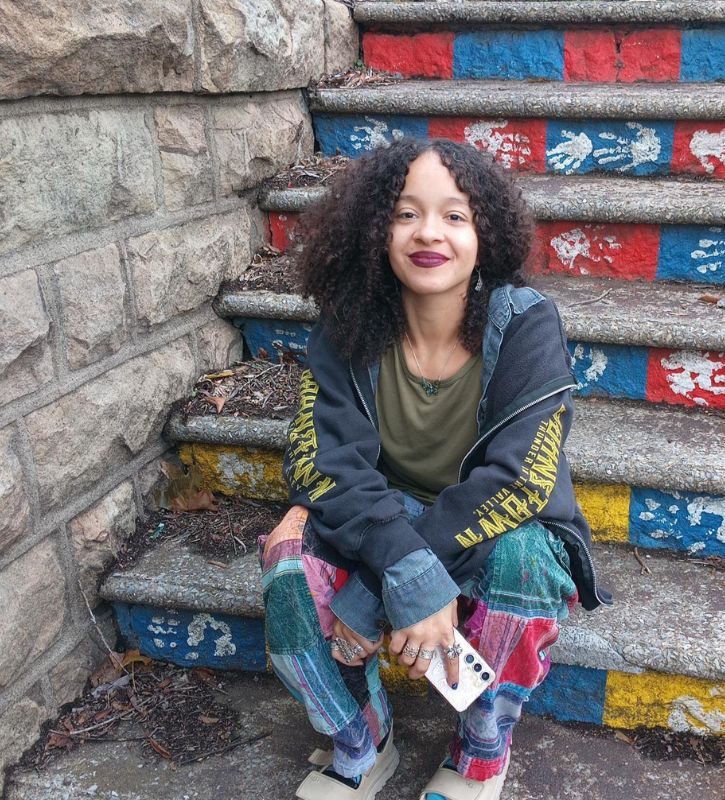 Photo of a mixed-race woman with shoulder-length dark brown curly hair sitting on painted stairs, wearing a gray zip up hoodie, patchwork, pants, and burgundy lipstick.