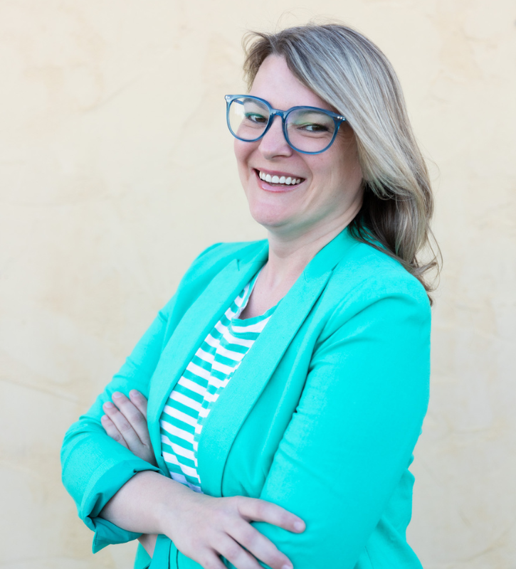 Aleshia is a white woman with past-should length blonde hair. Aleshia is wearing blue framed glasses, a teal blazer, and a teal and white striped shirt. She is smiling for the camera with her arms crossed in front of her.