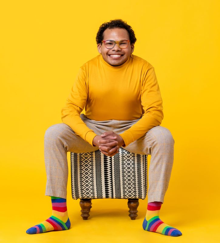 Headshot of Justin Tapp, a Black advocate with short hair wearing orange glasses, a yellow turtleneck, plaid pants, and rainbow-colored socks.