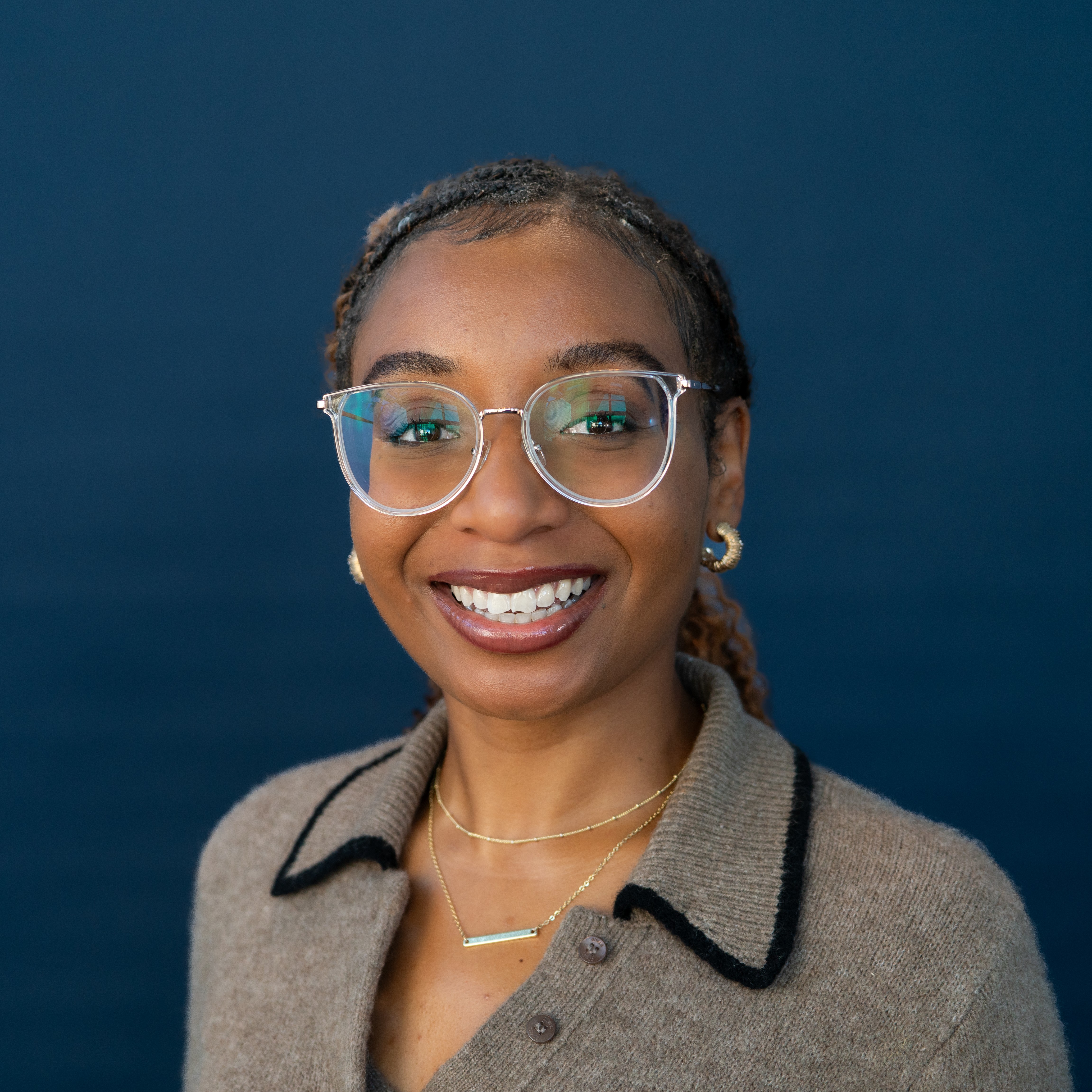April is smiling. April is a Black Woman with braids pulled into a ponytail. She wears glasses, a gray dres with a collar, and gold jewelry.