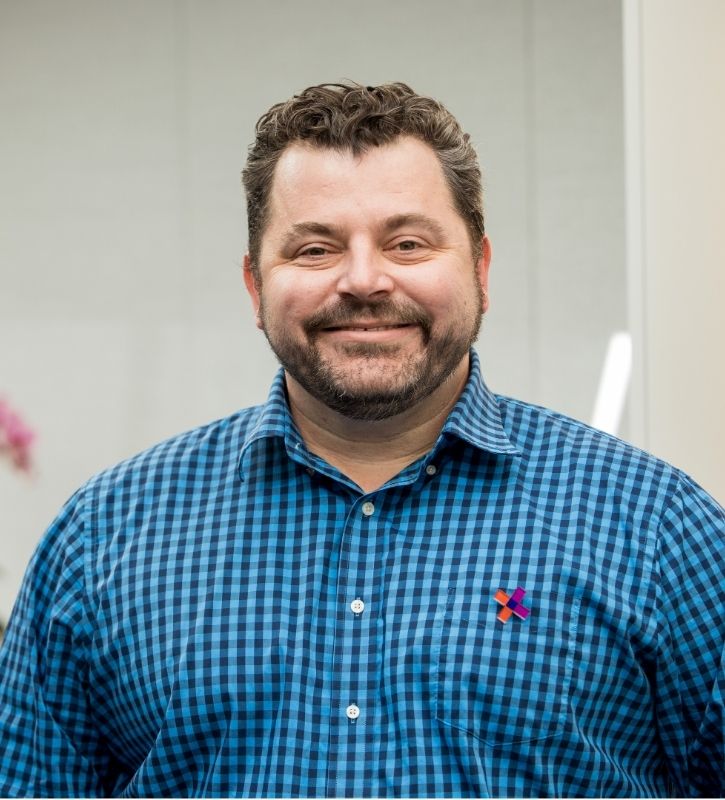 Jess Grainger is a white man with short brown, loose curly hair and gray and black facial hair wearing a blue checkered shirt .