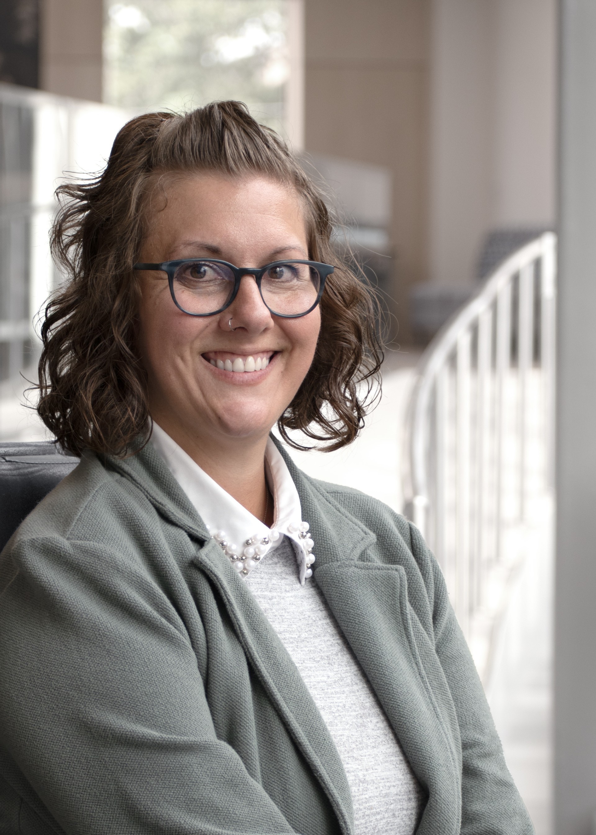 A White woman wearing a gray blazer, a collared shirt, and glasses. Christie is smiling with chin length loose curly hair.
