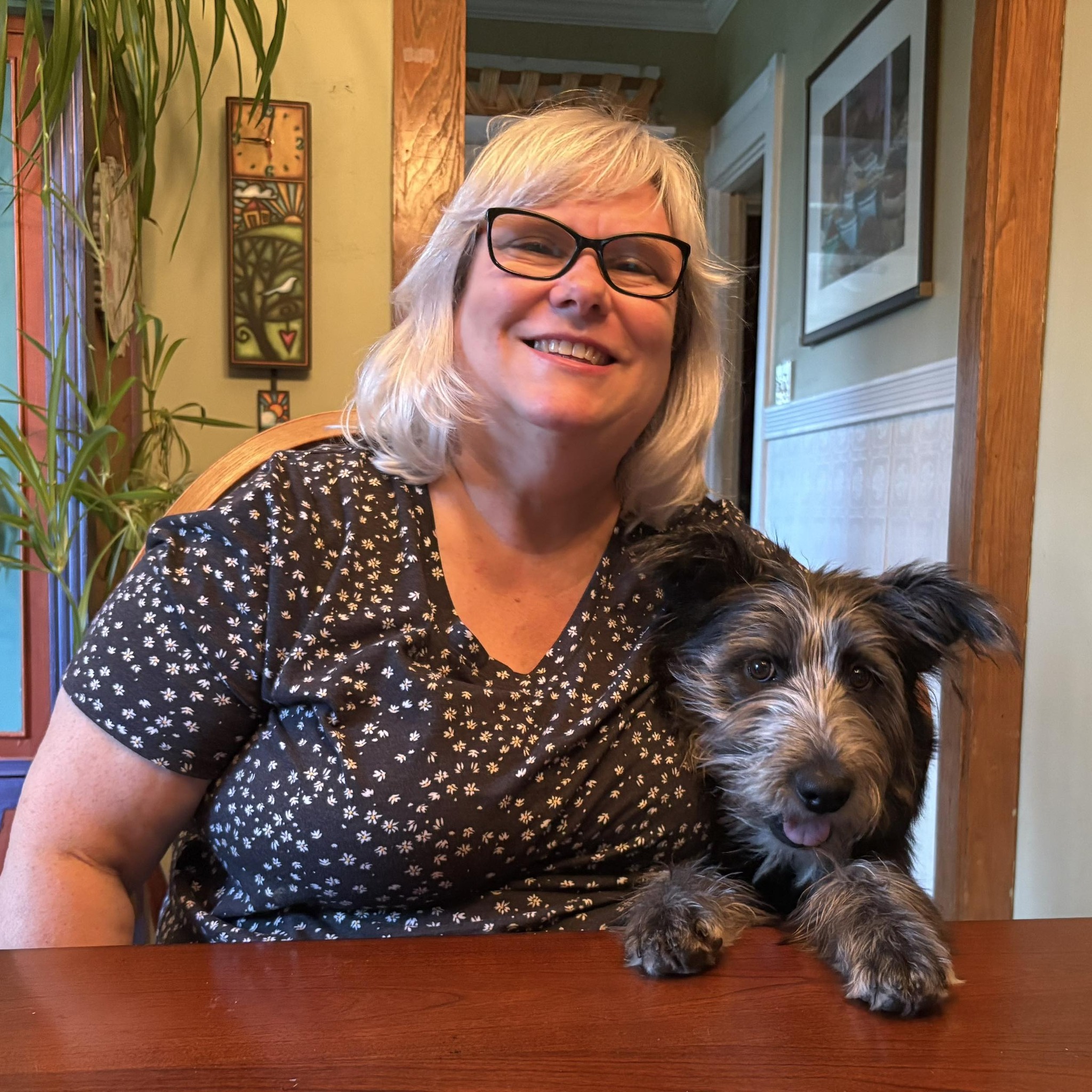 White woman with blonde shoulder-length hair and bangs. She is photographed with a dog wearing a Black polka dot blouse.