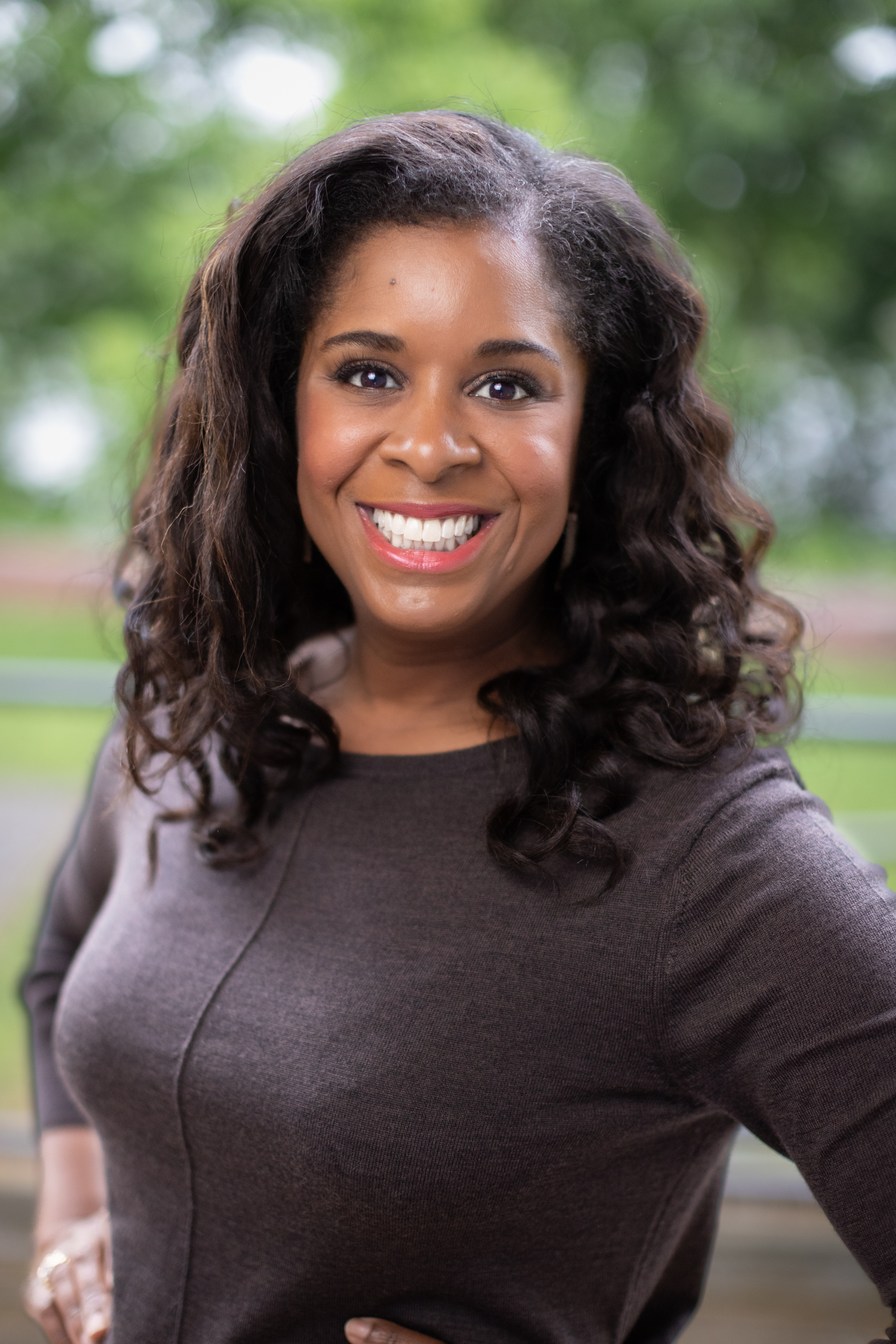 A Black woman with medium to dark complexion with Shoulder-length black hair. She is smiling and wearing a dark long-sleeves shirt.