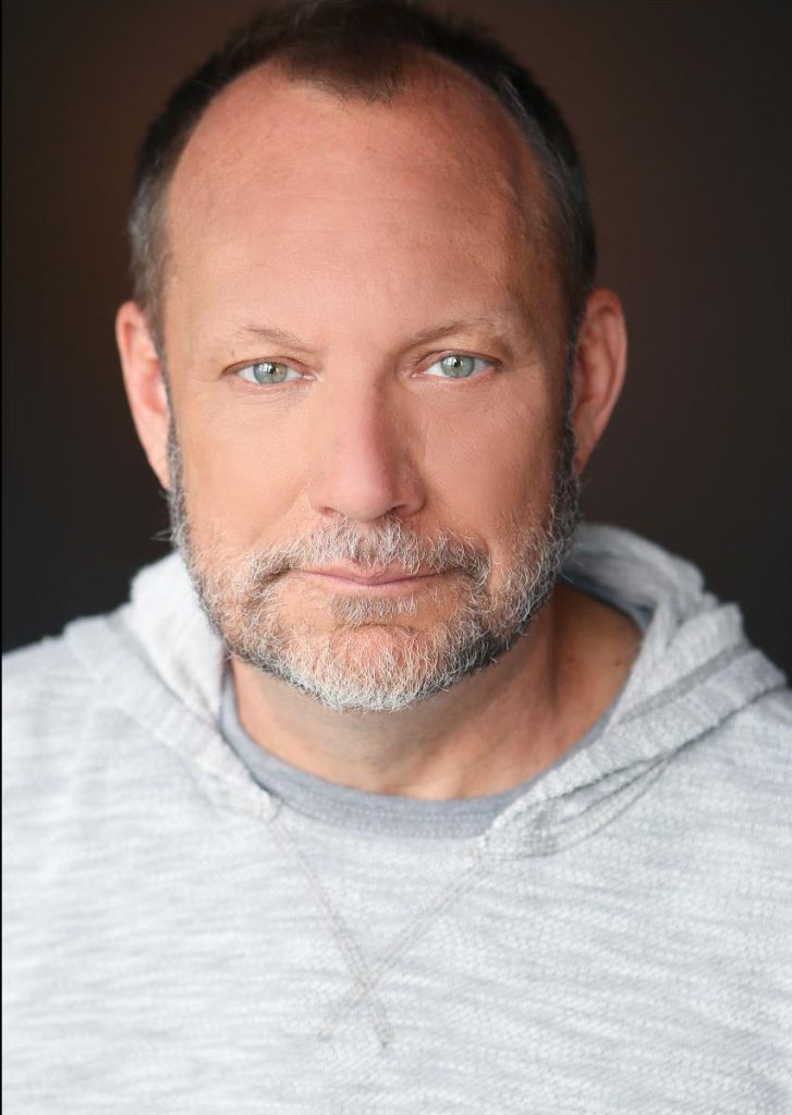 A white man with salt and pepper short hair and bear. He is wearing a hooded gray shirt.