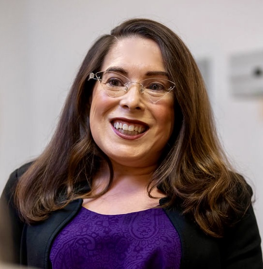 A White woman with chest length brunette hair wearing dark lipstick, glasses, and a black cardigan and dark gray shirt. 