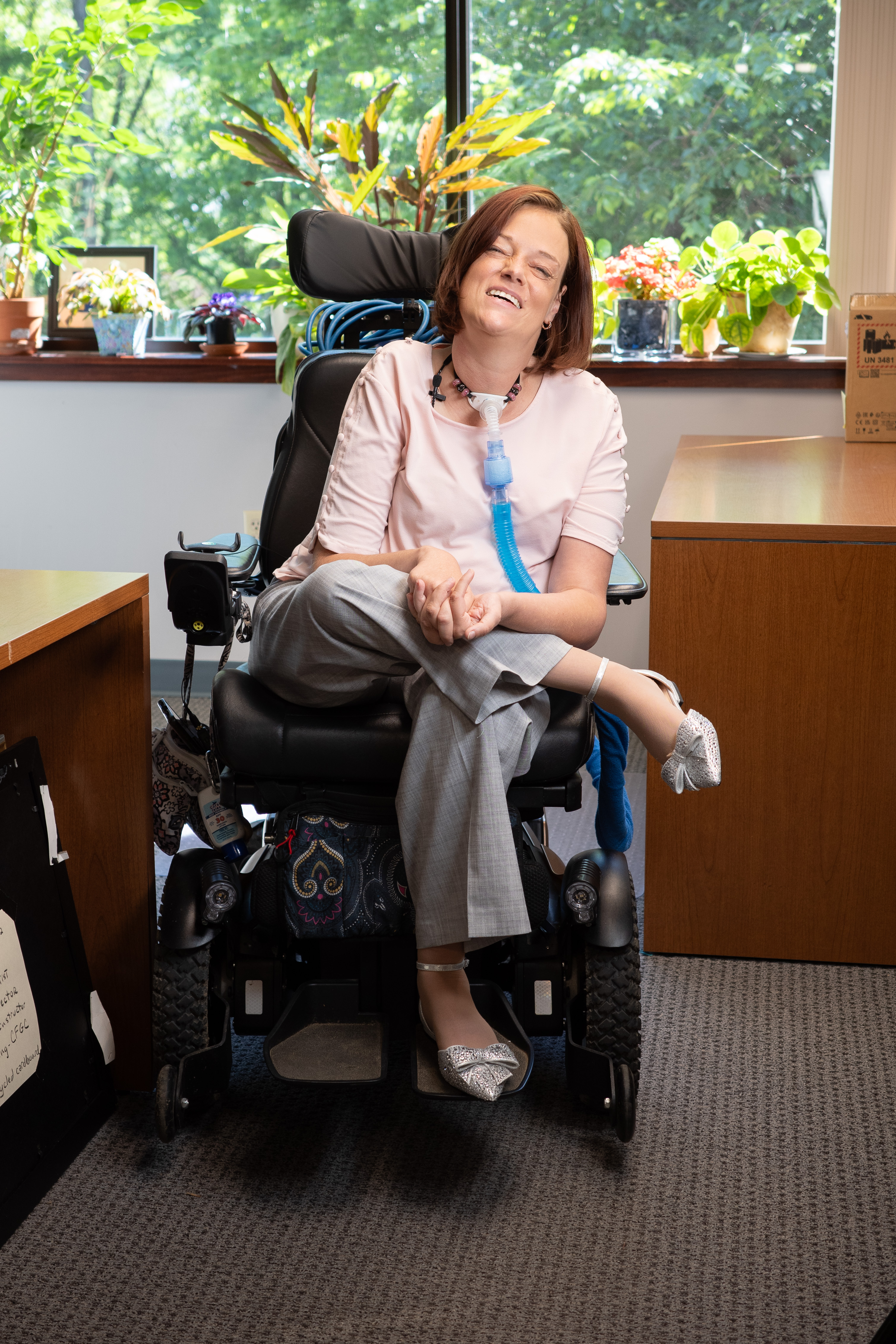 Josie is smiling and sitting in a power chair wearing. Josie is wearing a yellow blouse and is in an office setting.