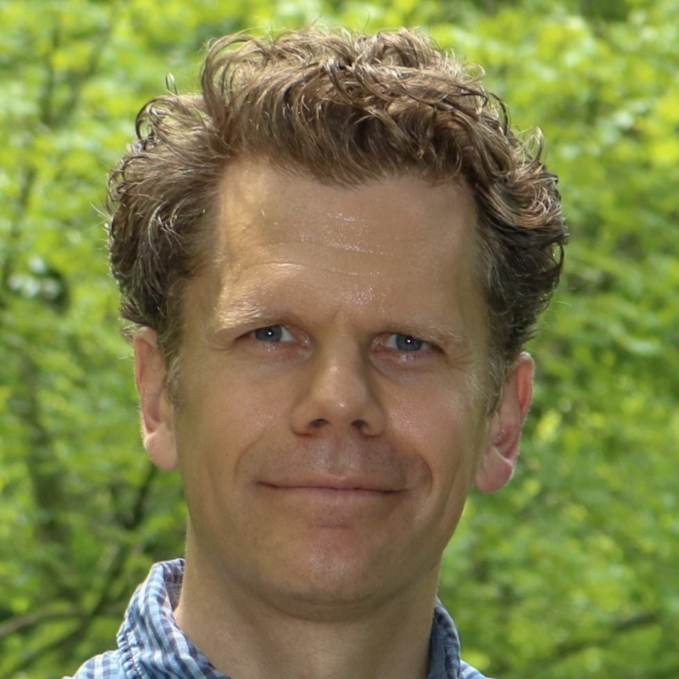 Craig is a White man with short brown hair and wearing a paid blue shirt. Craig is smiling in an outdoor setting.