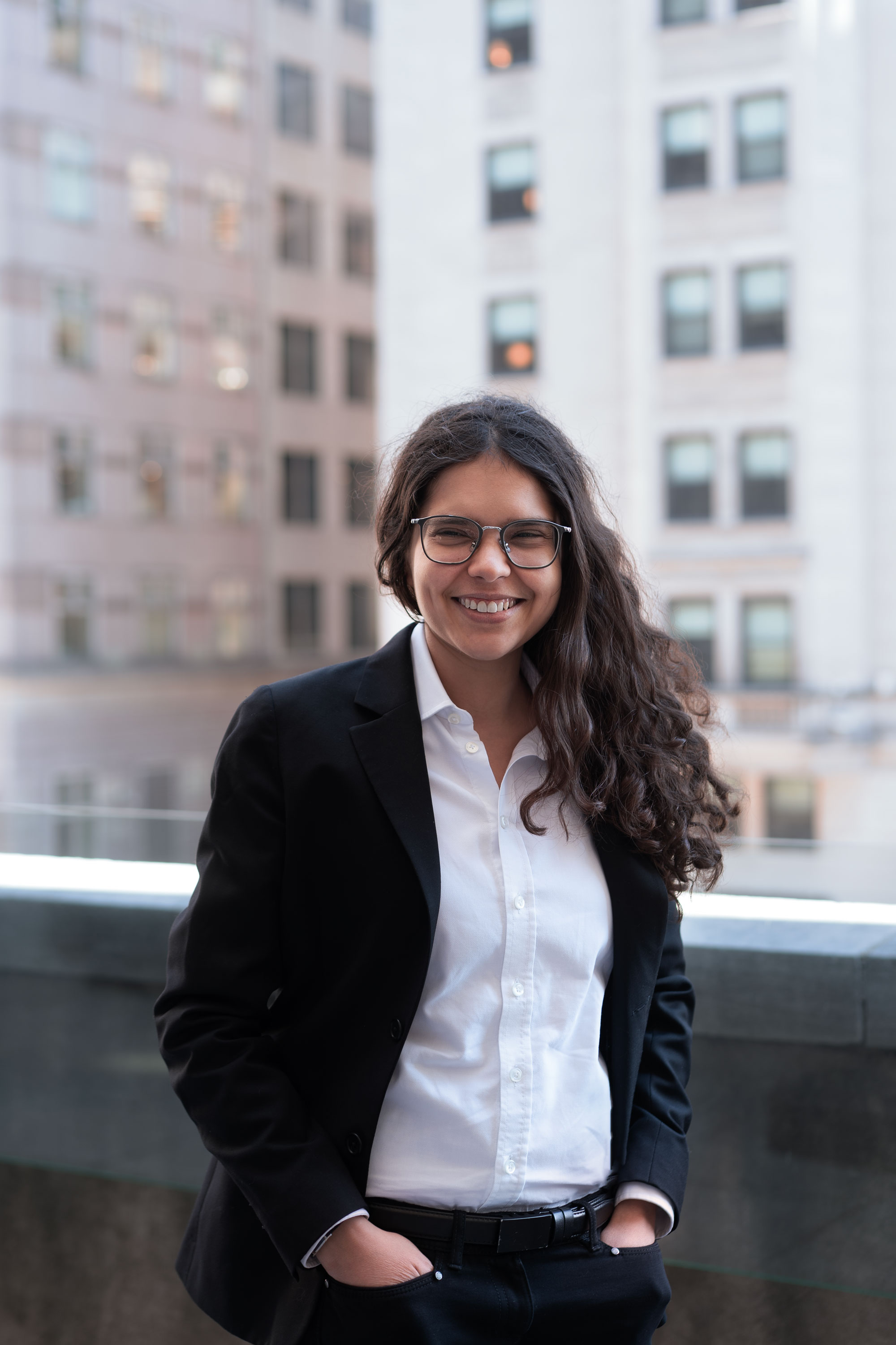 A person with long dark curly hair wearing a white button down with black blazer and dark wireframed glasses on a roftop of a building.