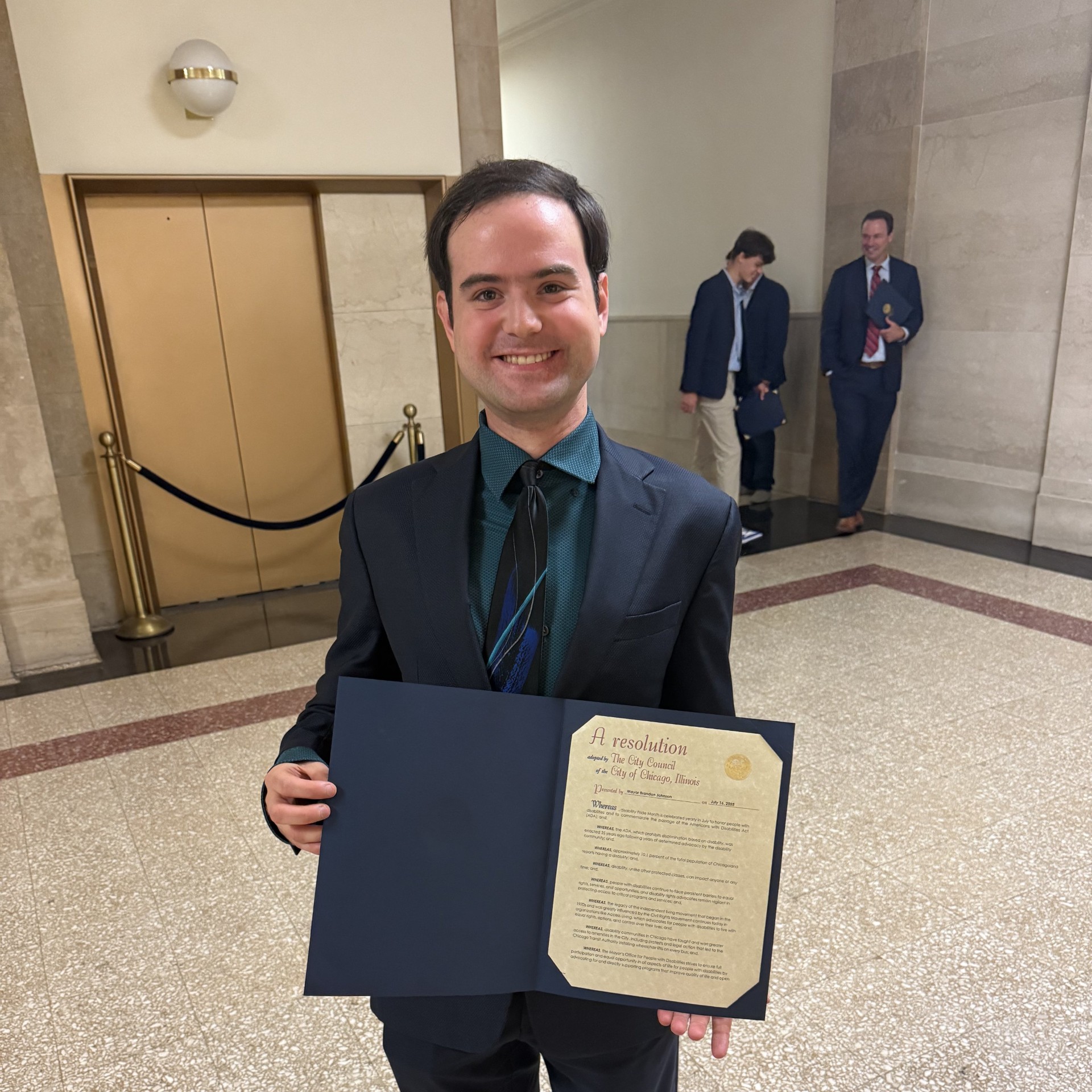 Eli. a white man with short brown parted on the right side,  is smiling and holding a siggned government declaration in a a government building. Eli is wearing a black suit with a dark blue shirt and blue striped tie.