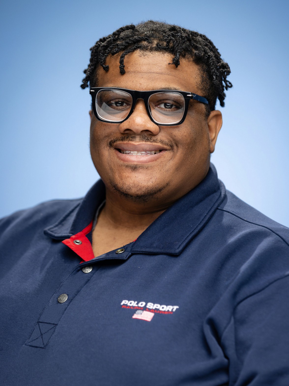 Timotheus is a Back man with short dreadocks and a UIC polo and is smiling against a bue background.