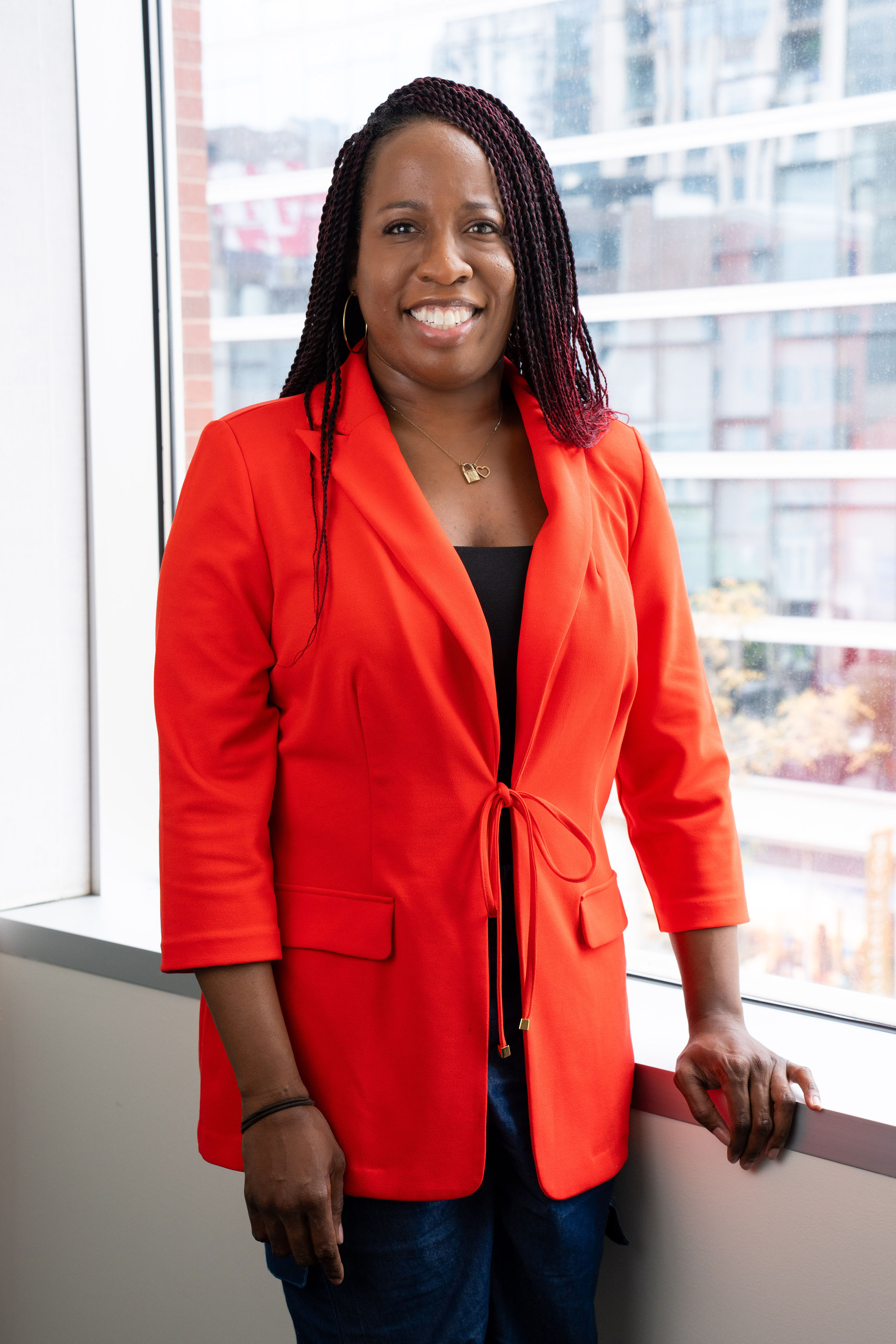 Candace, a Black woman with deep brown skinned shoulder length dark straight hair, is standing near an office window and is smiling. Candace is wearing a bright reddish orange blazer and black pants.