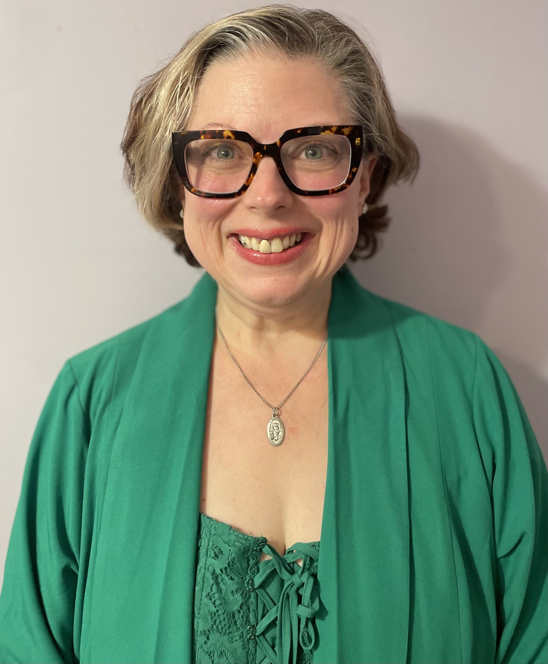 Cheryl, a white woman with chin-length blonde hair styled in loose waves and wearing thick framed glasses and a green cardigan, is smiling in an indoor setting.