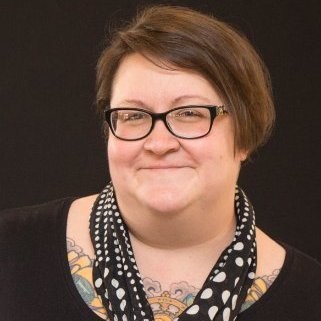 Jaime, a white woman readdish-brown hair combed over to her right ear, is wearing drak framed glasses and a dark t-shirt. Around Jaime’s neck is a black statement polka dot scarf and tattoos with various illustrations including a flower.