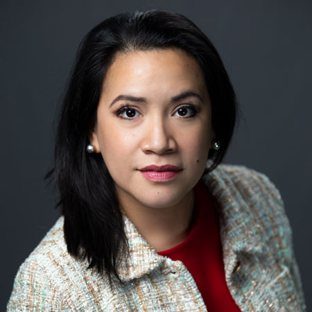 Susana is a Latina woman with light brown skin and long dark hair and pearl earrings, wearing a multicolored tweed jacket over a red top.