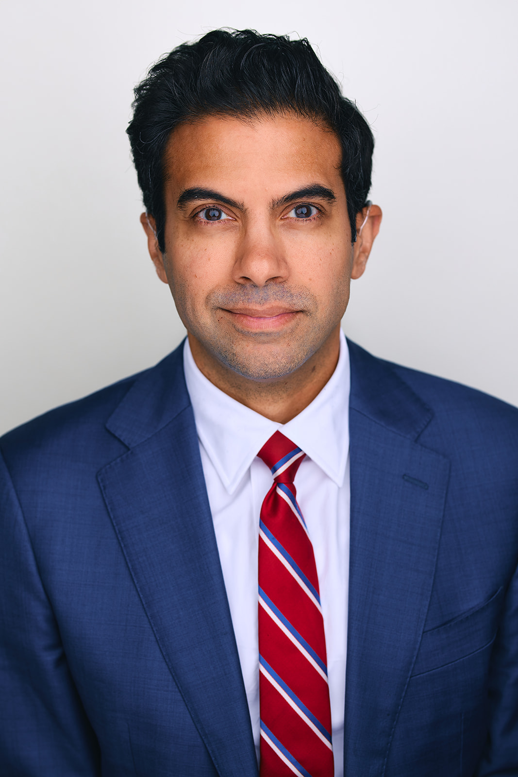 Richard is a man with brown skin and short black. He wears a white shirt and blue sportscoat with a gray red a striped tie.