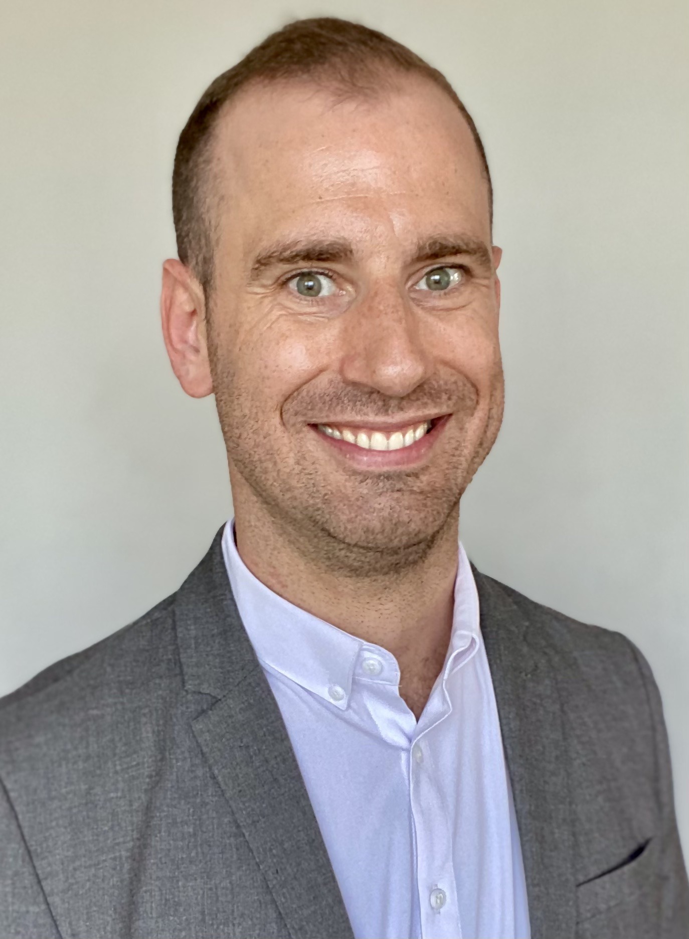 Brian, a white man with short buzzed dark brown hair wearing a gray blazer, white shirt, and light blue tie, is smiling against a gray background.