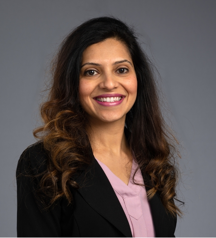 Kruti, a brown woman with long dark curly hair wearing a black blazer and rose color blouse, is stands smiling in front of a gray background.