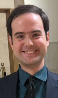 Eli, a white man with short brown parted on the right side,  is smiling in a government building. Eli is wearing a black suit with a dark blue shirt and blue striped tie.