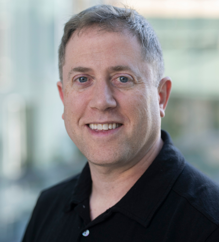 Andrew is wearing a black collared shirt, and smiling against a blurred out background. 