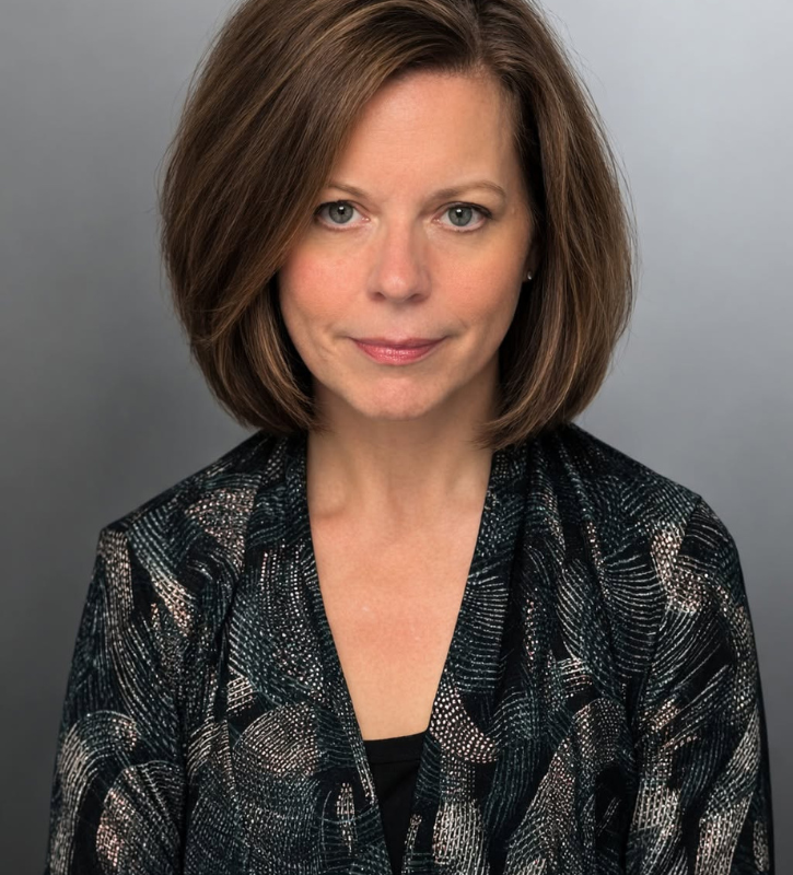 Carla Anderton is a white person with brown hair cut into a bob and is smiling. Carla is wearing a patterned blouse and blazer.
