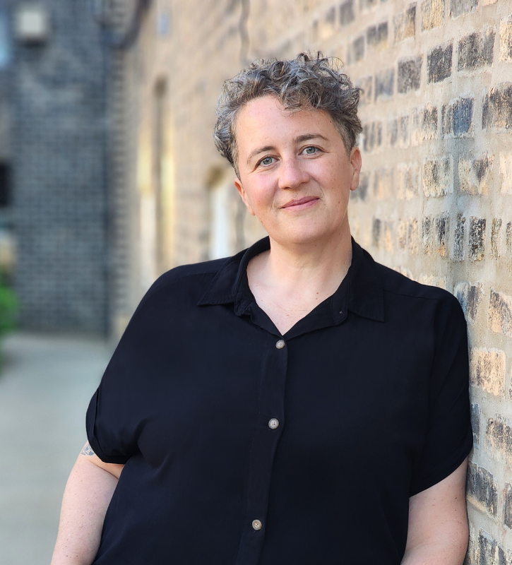 A headshot of Cassidy Dimon, a white queer woman with short curly brown and gray speckled hair leans against a brick wall wearing a black button up shirt.