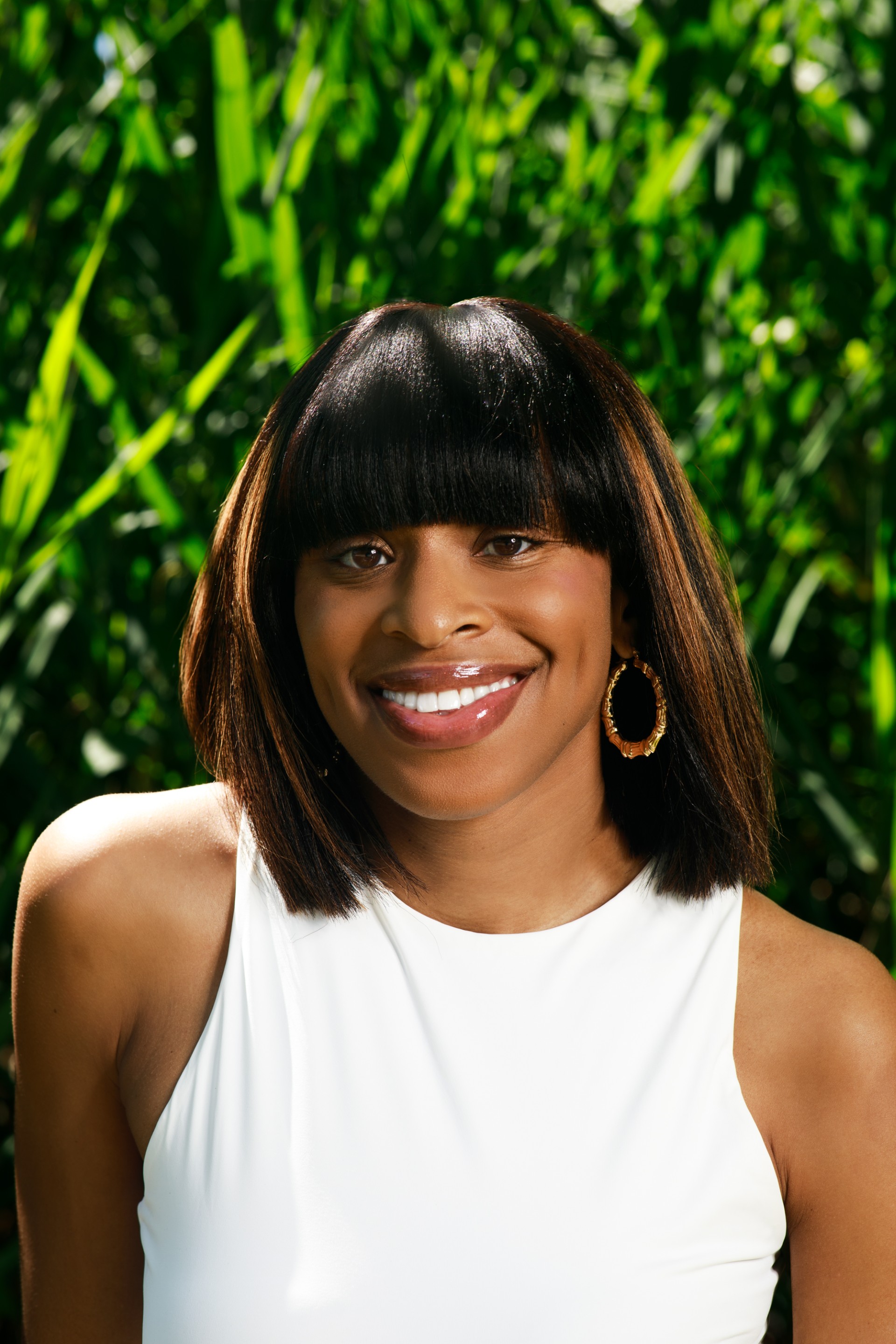 Gizelle, a black person with shoulder-length black hair and bangs, is smiling and wearing a white top.