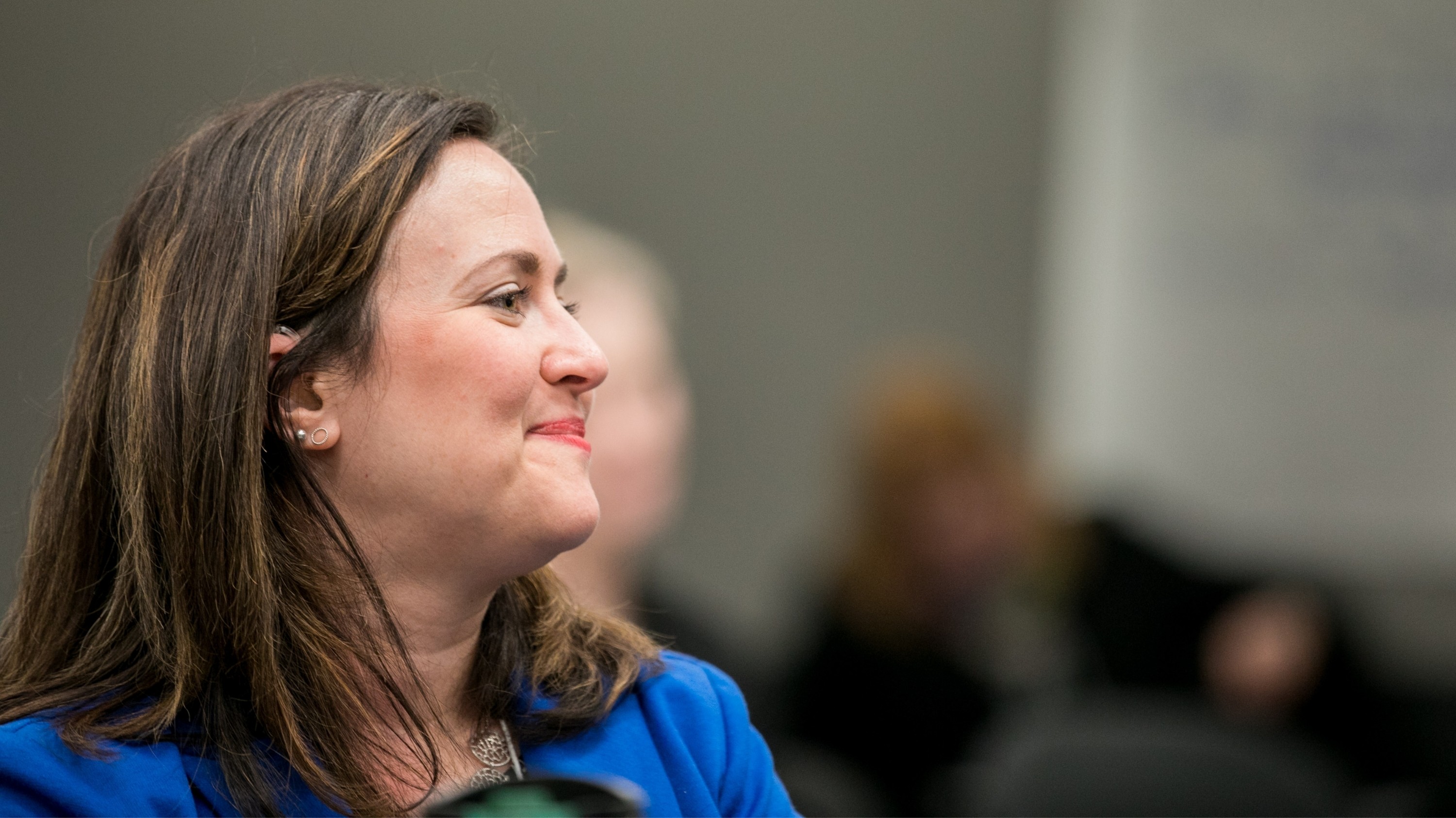 A woman with long brown hair, medium-light toned skin, a blue dress, and a serene expression looks off to the side.