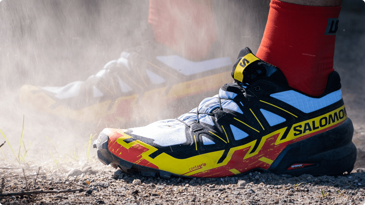 Close up of a Salomon running shoe in the colors red, yellow, white, and black. The person's feet are surrounded in a cloud of dust. 