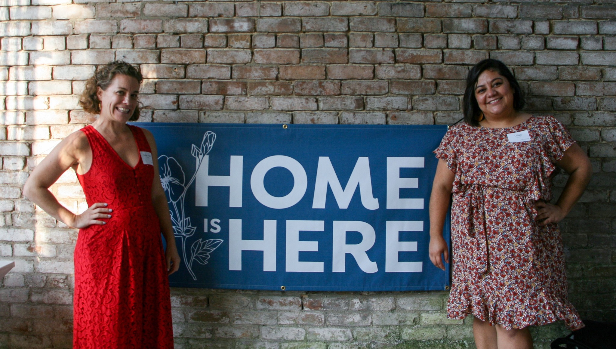 Two women in flowy dresses standing in from of a Home is Here banner, smiling brightly.