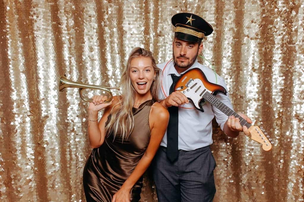 Pareja posando frente a un fondo dorado brillante con atrezzo divertido de Selfriends Fotomatón: ella sostiene una trompeta y él una guitarra de juguete con gorra de capitán.