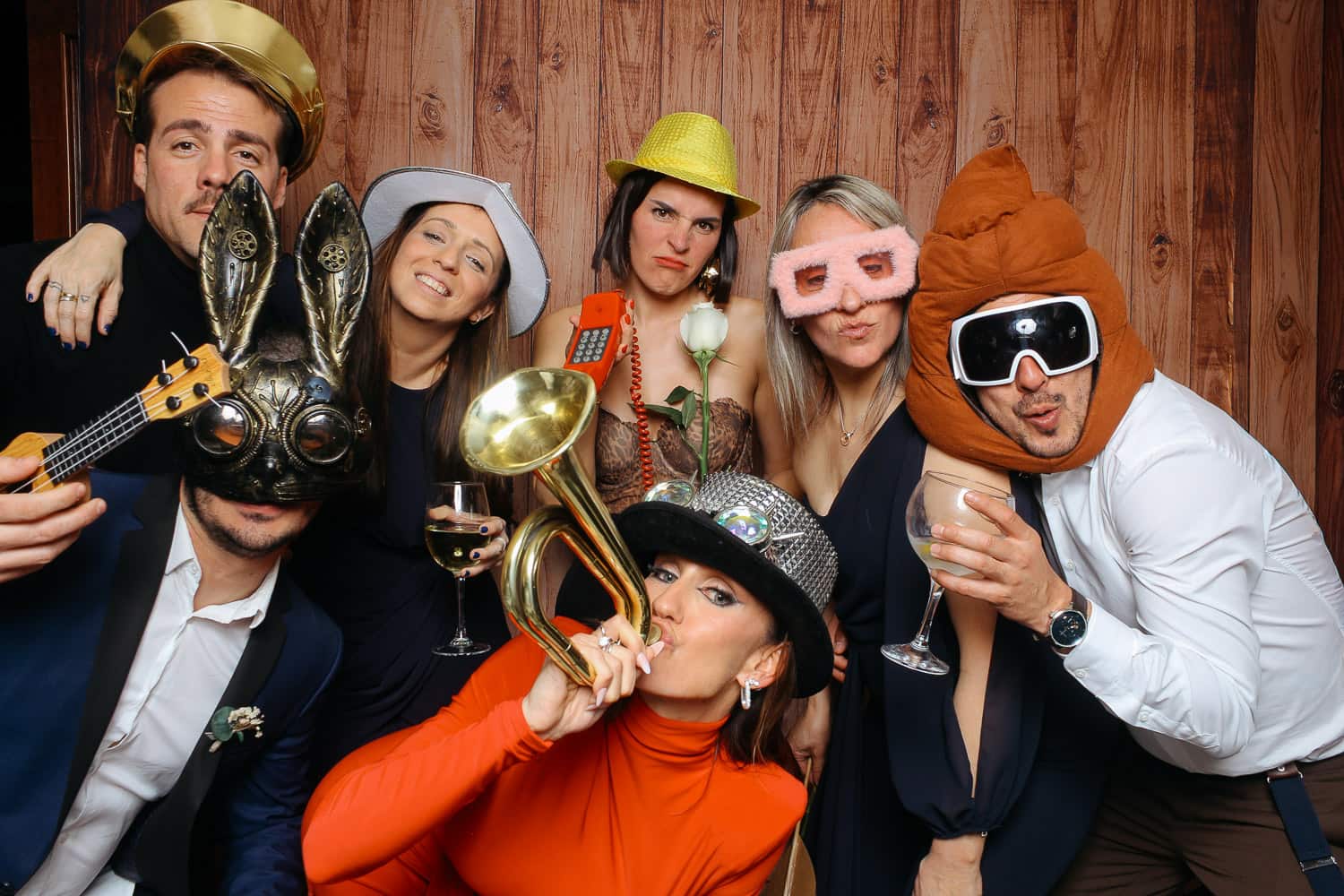Grupo de personas posando divertidas con atrezzo original de Selfriends Fotomatón: sombreros, gafas gigantes, máscaras y accesorios coloridos frente a fondo de madera.
