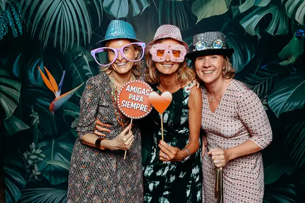 Tres amigas posando con accesorios divertidos en el fotomatón Selfriends durante una boda en Barcelona, con fondo tropical y cartel “Amigas para siempre”, capturando momentos reales y espontáneos.