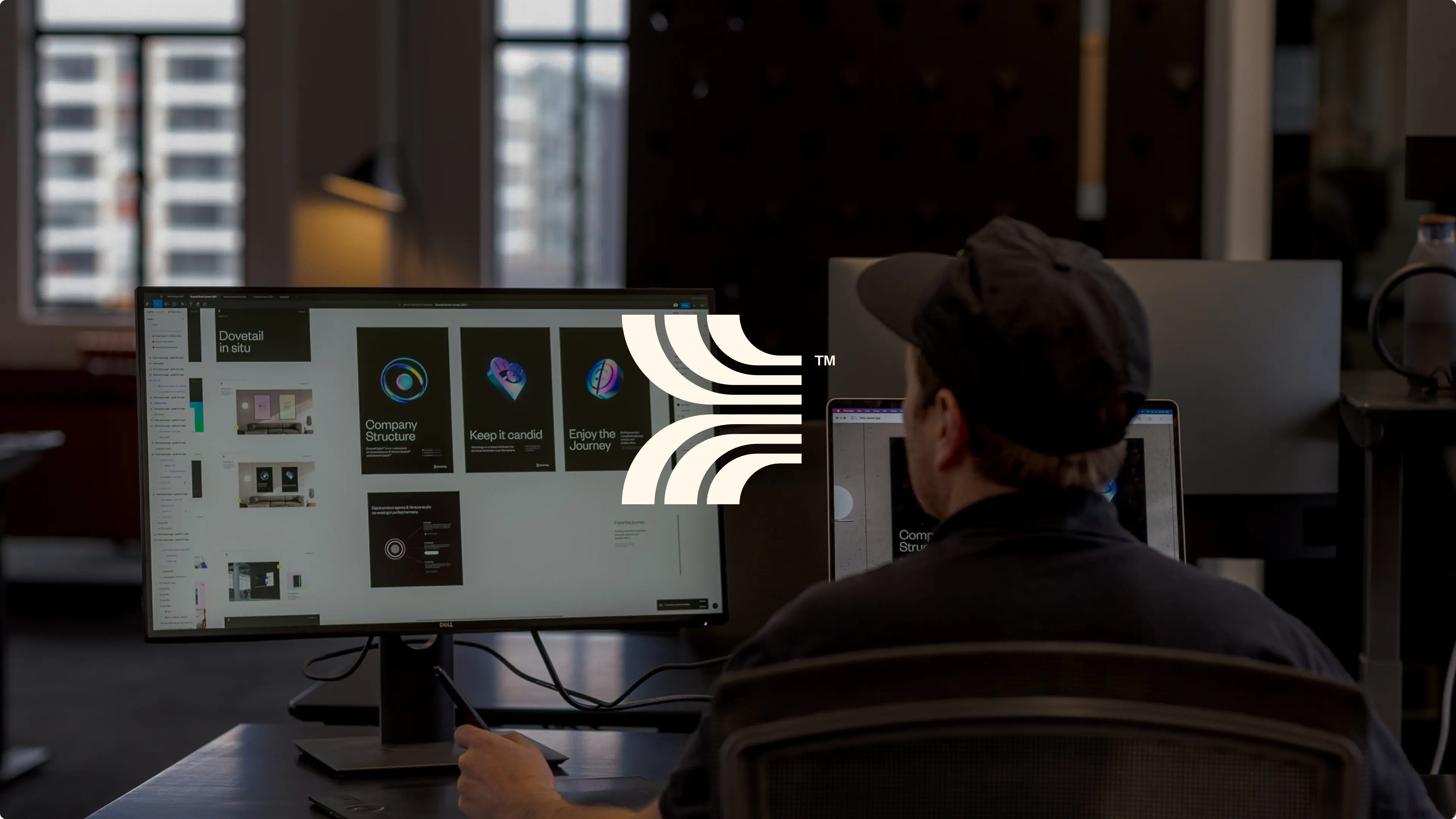 Person wearing a black cap working on dual monitors displaying design projects in a dimly lit office.